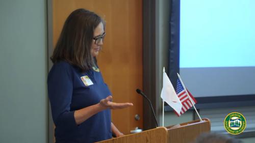 A women stands behind a podium with Japanese and American flags